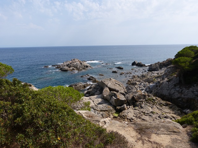 Vista costanera amb formacions rocoses, mar en calma, vegetació i cel parcialment ennuvolat