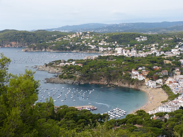 Vista des del Far de Sant Sebastià amb port ple de barques, turons verds i edificis residencials