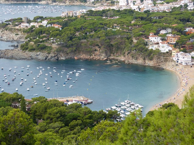 Vista des del Far de Sant Sebastià amb badia plena de barques, platja i turons verds amb edificis