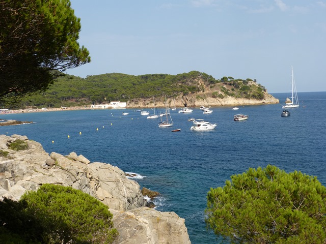 Vista costanera amb roques, vegetació, velers ancorats i una illa o península al fons