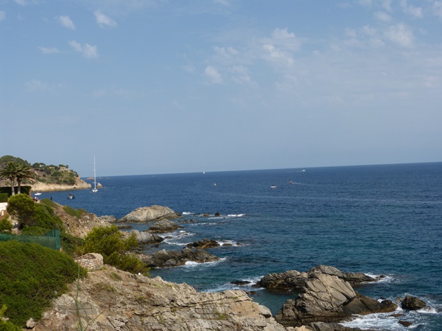 Vista costanera amb penya-segats, vegetació, velers sobre el mar i cel clar amb núvols suaus