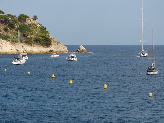 Vista costanera amb velers sobre mar blau, boies grogues, roques i vegetació lateral