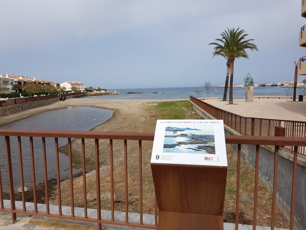 Porta d’entrada al Cap de Creus amb panell informatiu, palmera i edificis costaners al fons