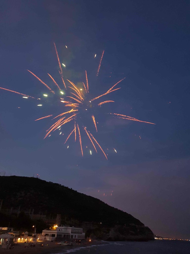 Focs artificials sobre el mar en una nit festiva amb edificis i turons al fons
