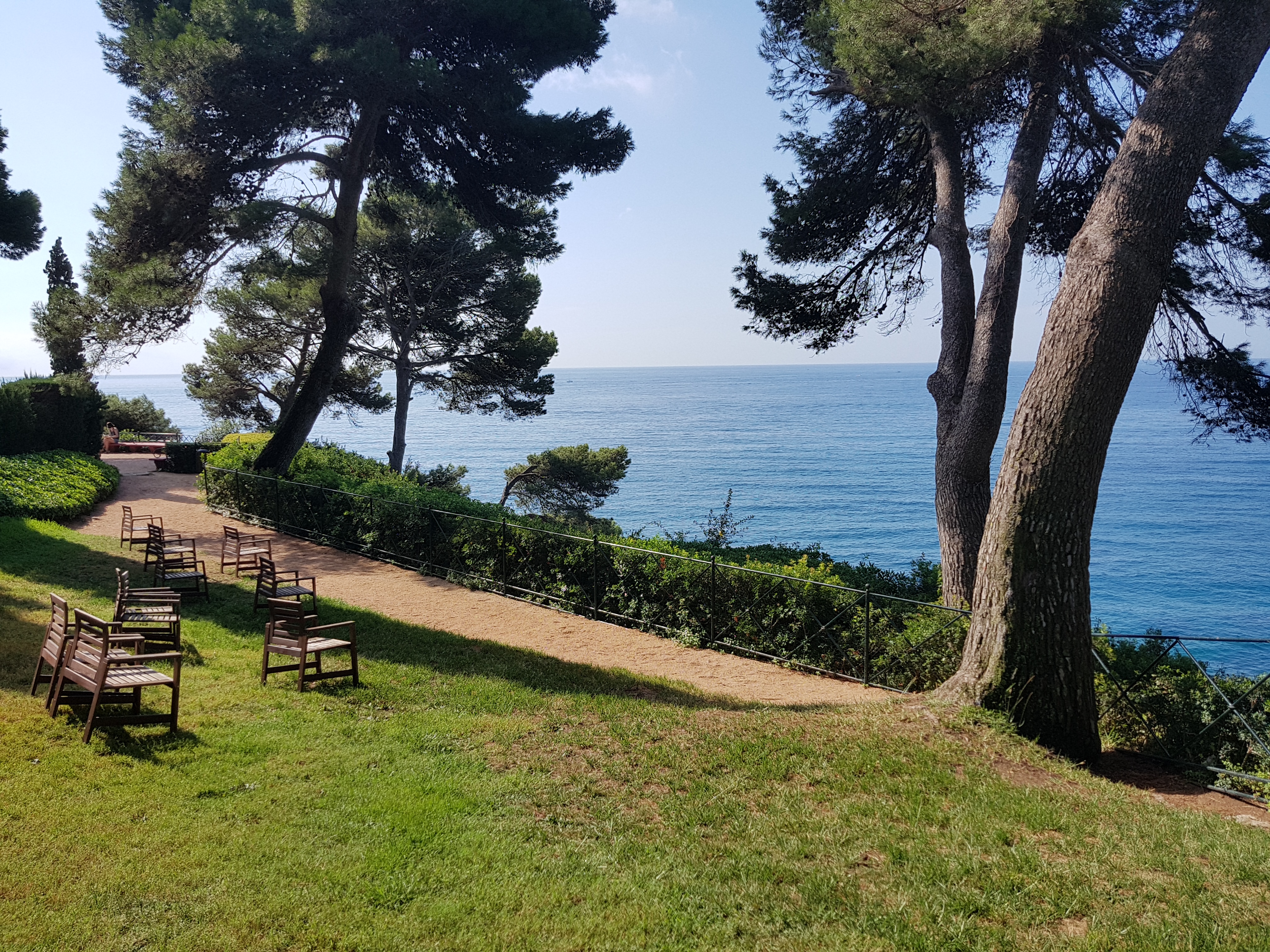 Taules i cadires de fusta entre pins amb vista al mar als Jardins de Santa Clotilde