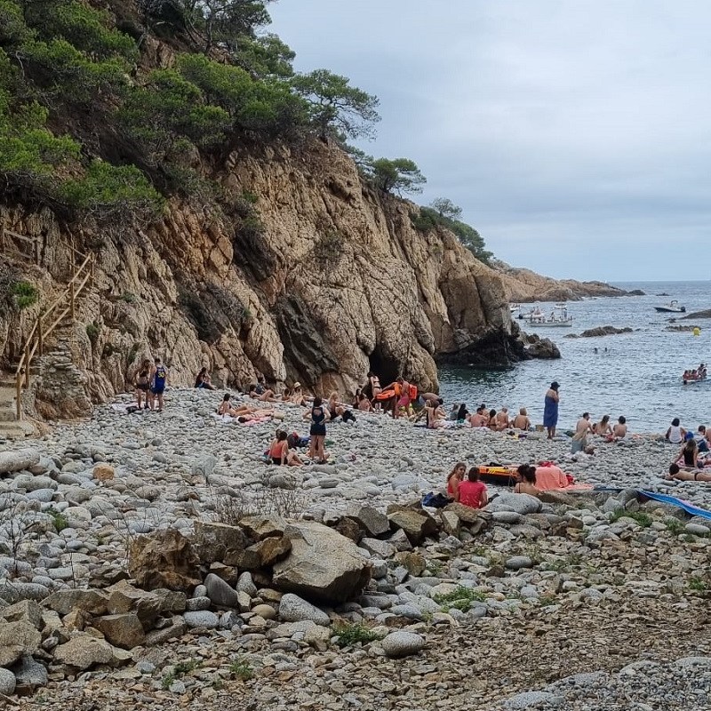 Cala Pedrosa amb roques, persones a la platja i barques al mar