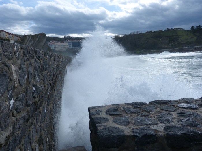 Onada trencant contra mur de pedra amb muntanyes verdes i cel gris al fons