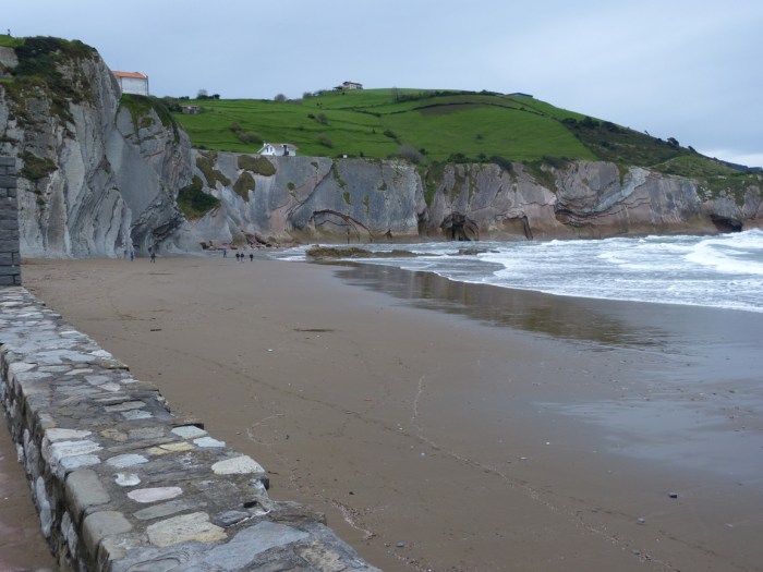 Platja amb mur de pedra, penya-segats verds i mar Cantàbric sota cel gris