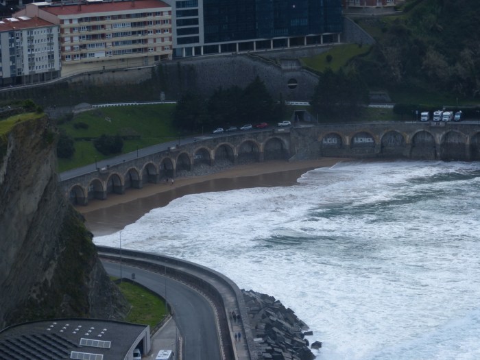 Onades trencant contra costa amb arcs, carretera i edificis al fons