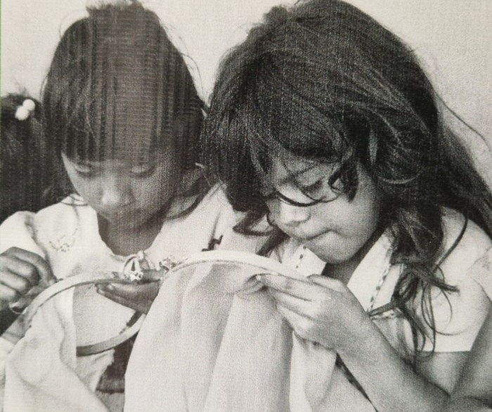 Fotografia en blanc i negre que mostra dues nenes  immerses en una activitat de brodat. Una observa mentre l’altra cus amb fil i agulla sobre tela. L’escena transmet concentració, aprenentatge i transmissió cultural.