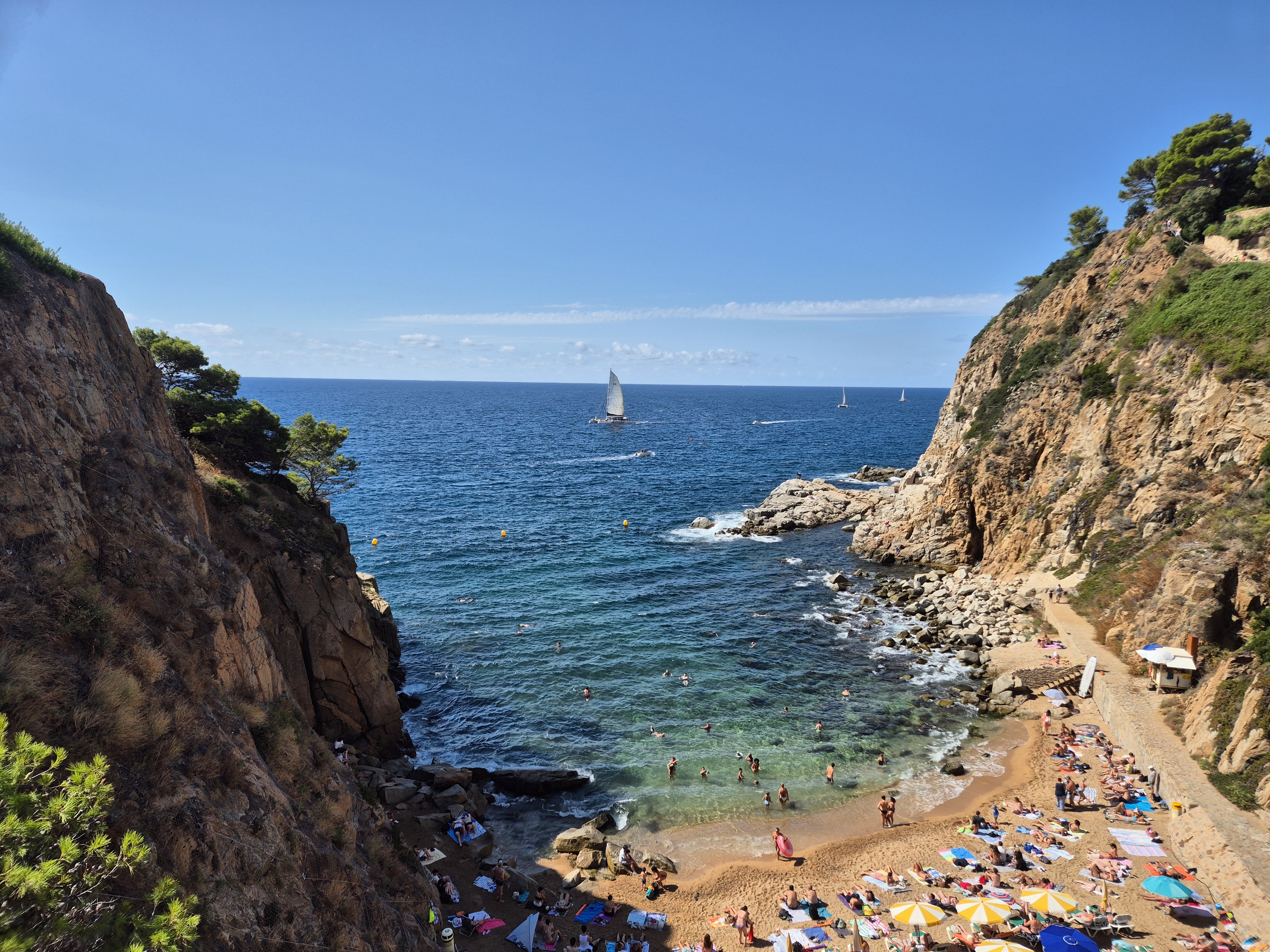 Vista d’una caleta amb banyistes, penya-segats rocosos i velers al fons