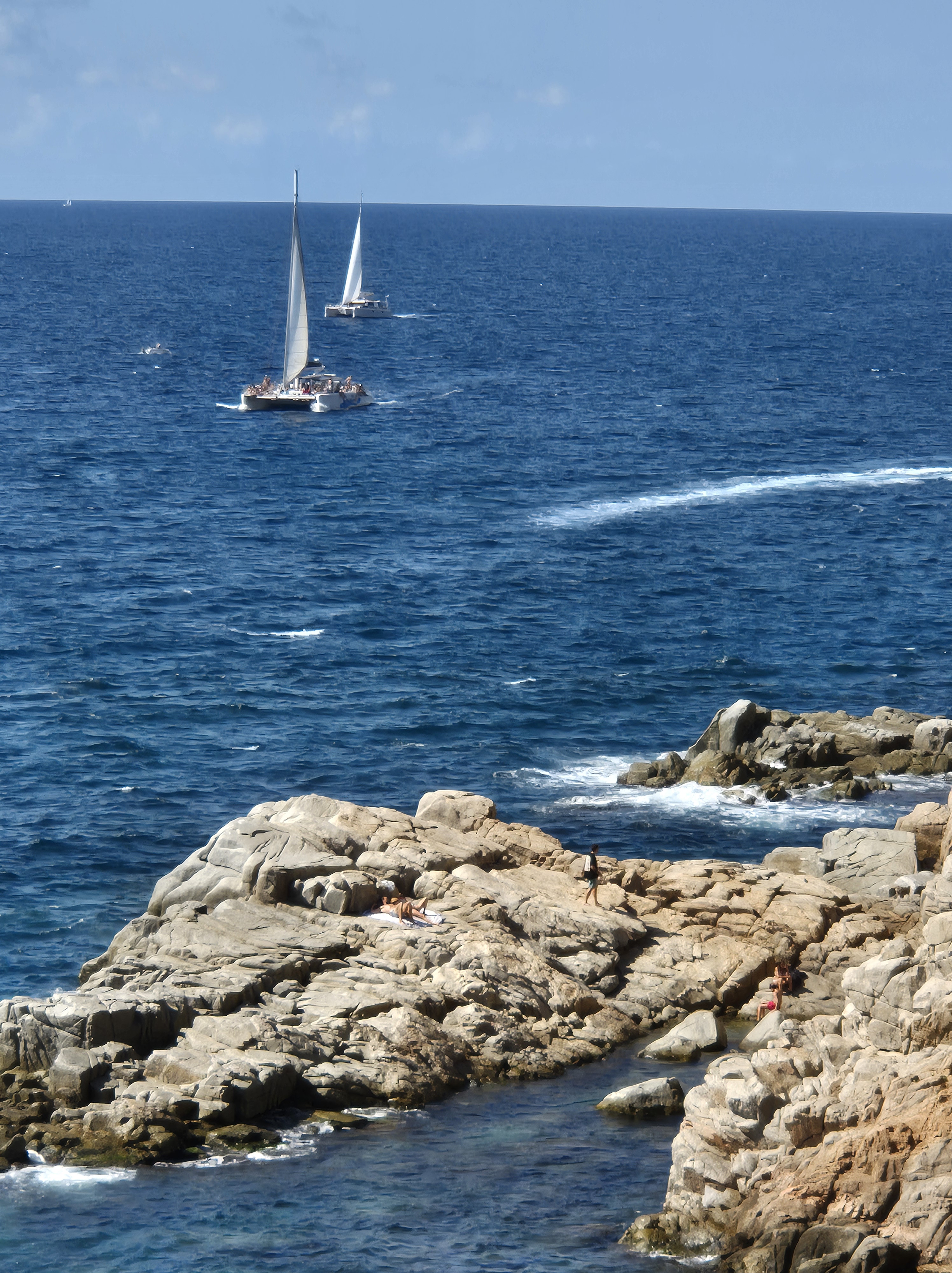 Persona asseguda sobre roques amb vista al mar i dues veles navegant