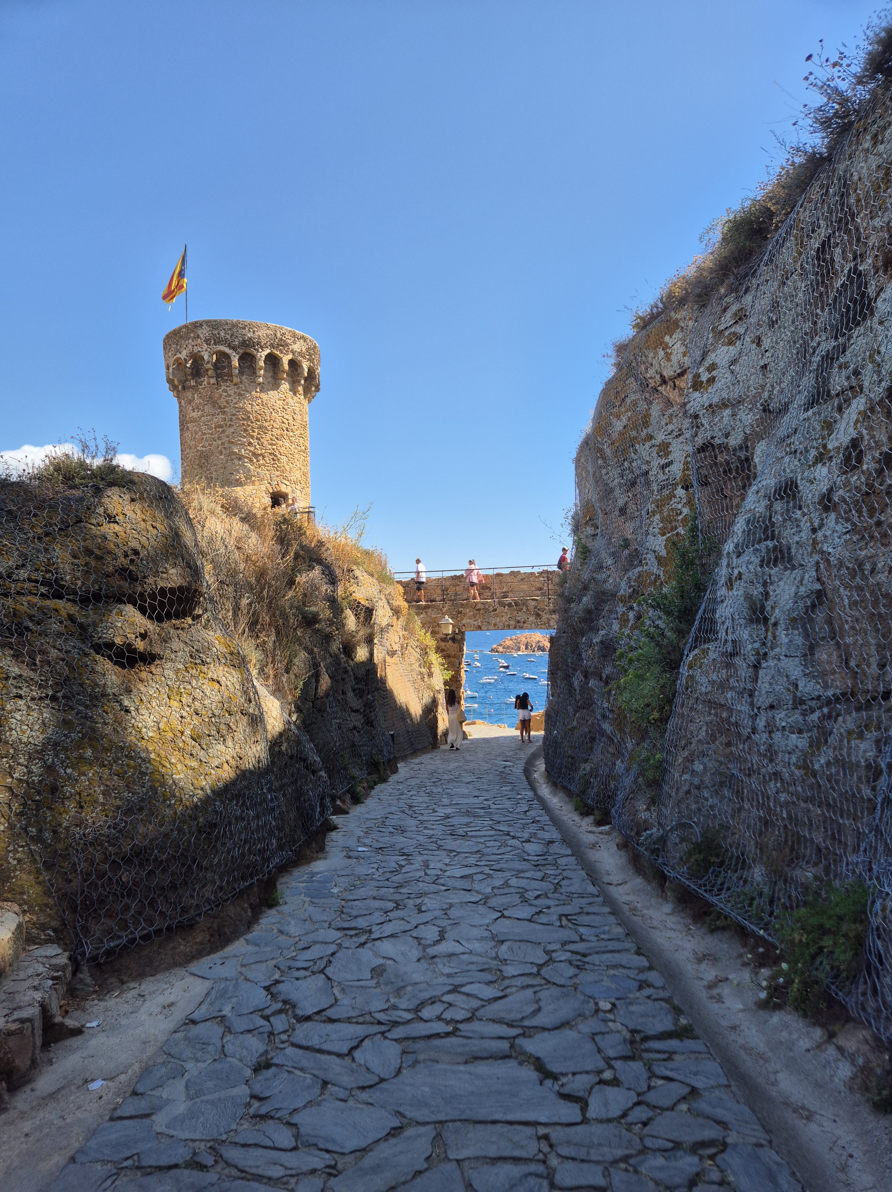 Camí empedrat entre roques que condueix a una torre medieval amb bandera, prop del mar