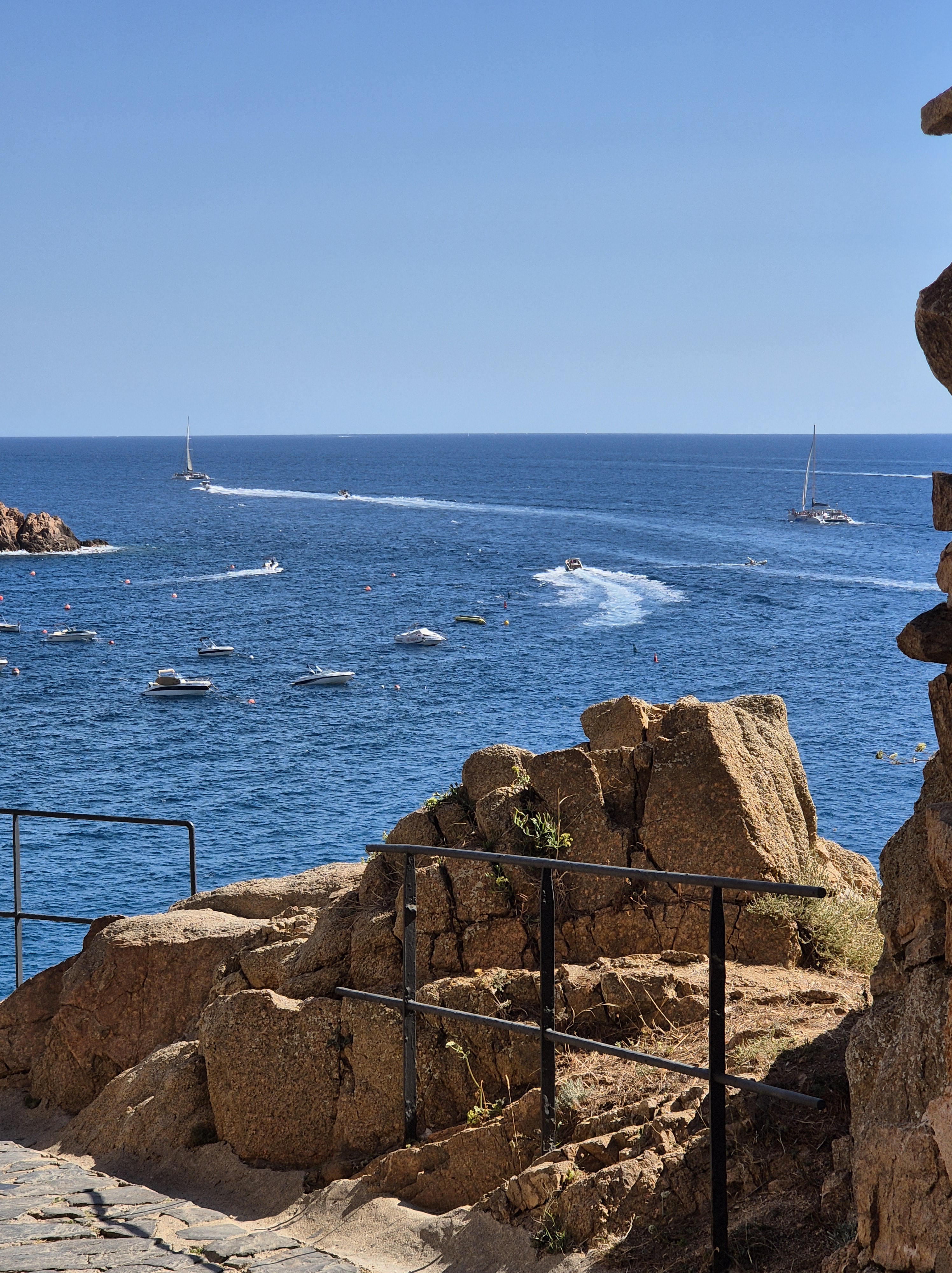 Vista des d’un camí rocallós amb barana, amb el mar blau i embarcacions al fons